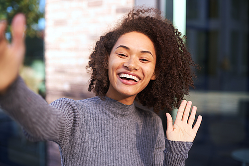 Attractive African American woman smiling a toothy smile, waving hello with her hand, expressing positive emotions while looking camera on a smartphone.