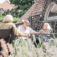 Bild zeigt einen Pfleger mit zwei älteren Frauen im Rollstuhl in einem Garten. 