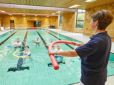 Eine Therapeutin steht vor einem Schwimmbecken und leitet ein Aquatraining an