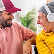 Begeisterter Mann mit Behinderung hört mit Kopfhörern Musik zusammen mit einem Freund.
