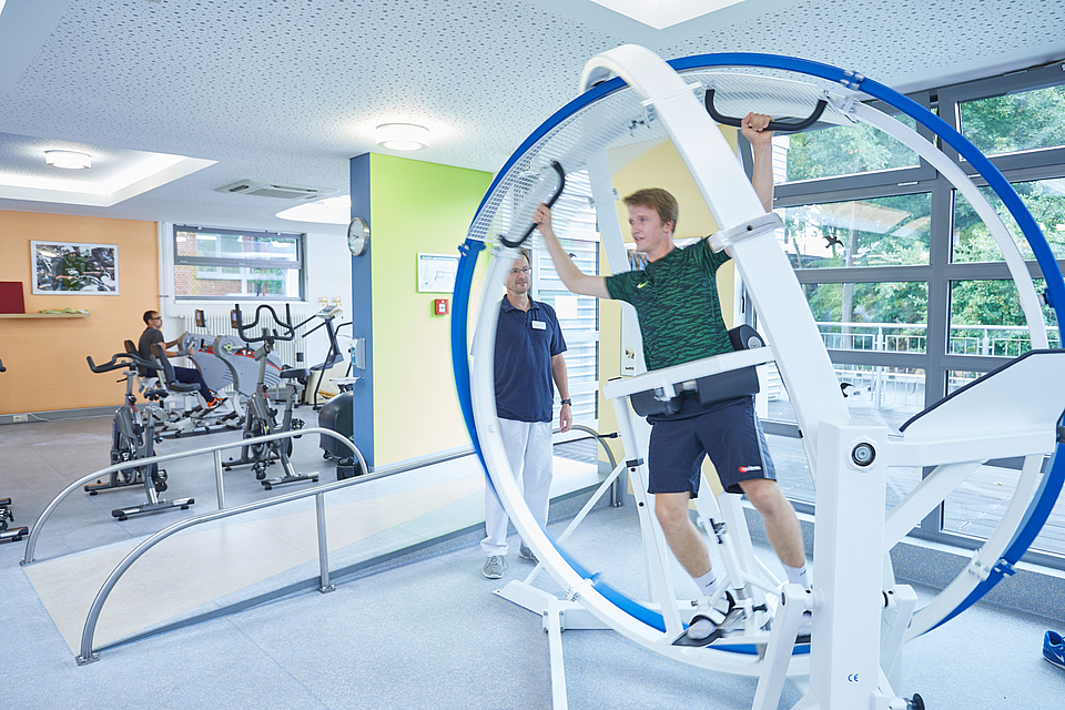 Ein Patient befindet sich im Spacecurl, einem modernen Therapiegerät zur Verbesserung der Bewegungskoordination und Muskelkontrolle. Der Patient wird von einem Therapeuten aufmerksam beobachtet, der ihm Anweisungen gibt und die Übung begleitet.